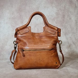 Peut inclure: Un sac &agrave; main en cuir marron avec une poign&eacute;e sup&eacute;rieure et une bandouli&egrave;re amovible. Le sac a une poche zipp&eacute;e &agrave; l'avant et un petit d&eacute;tail m&eacute;tallique. Le cuir a un aspect vieilli.
