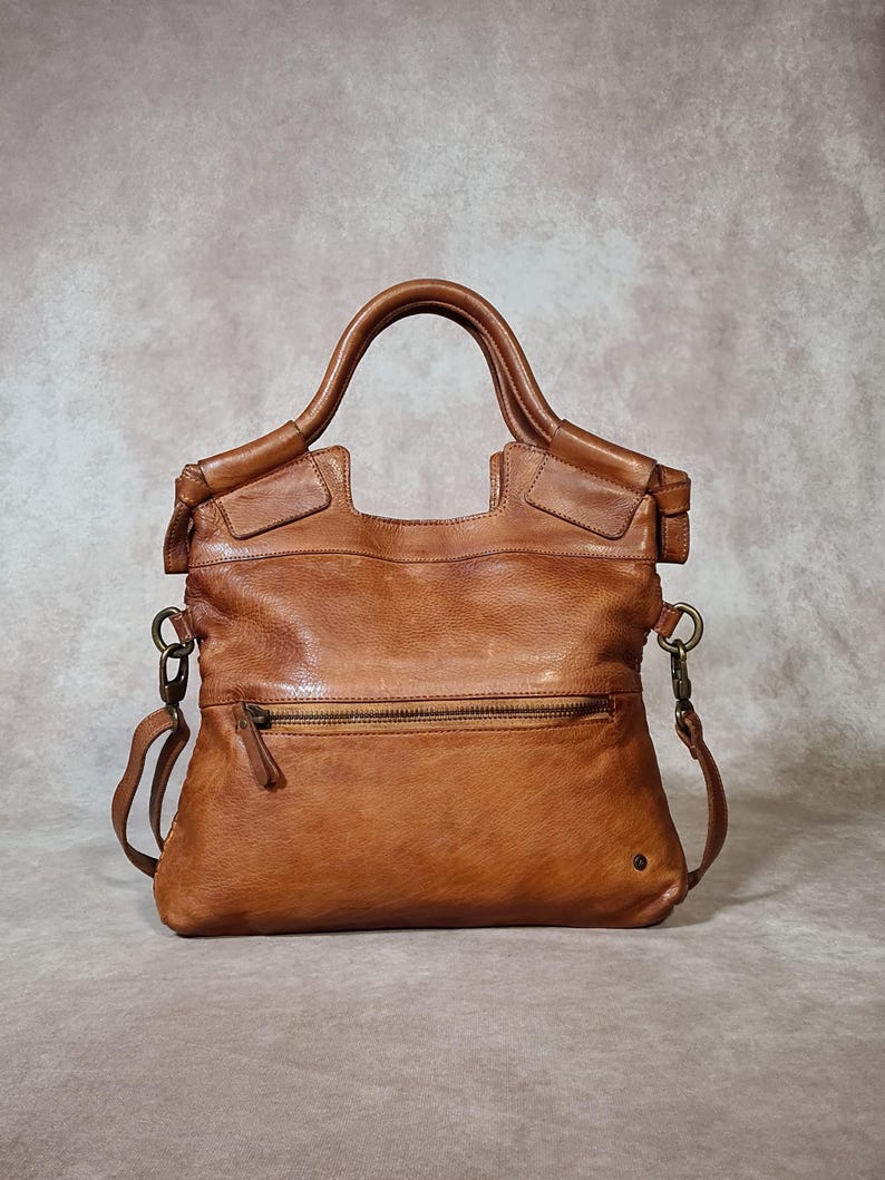 Peut inclure: Un sac &agrave; main en cuir marron avec une poign&eacute;e sup&eacute;rieure et une bandouli&egrave;re amovible. Le sac a une poche zipp&eacute;e &agrave; l'avant et un petit d&eacute;tail m&eacute;tallique. Le cuir a un aspect vieilli.