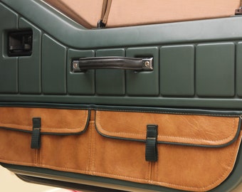 May include: A green car door interior with a brown leather storage pocket with a black strap. The door has a black handle and a silver accent.