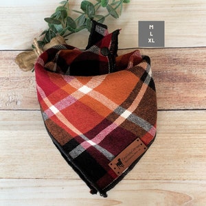 May include: A plaid dog bandana in shades of red, orange, white, and black. The bandana is triangular and features a leather tag with a dog silhouette and text. Sizes M and XL are indicated. The bandana is displayed on a wooden surface.