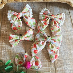 May include: A collection of fabric hair bows in various sizes. The bows feature a strawberry and floral pattern on a light background. Some bows have white lace trim. The bows are arranged on a wooden surface.