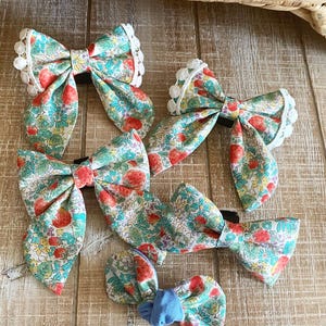 May include: A collection of fabric hair bows with a strawberry and floral pattern. The bows are in various sizes and feature a mix of red strawberries, green leaves, and blue flowers on a cream background. Some bows have white lace trim.