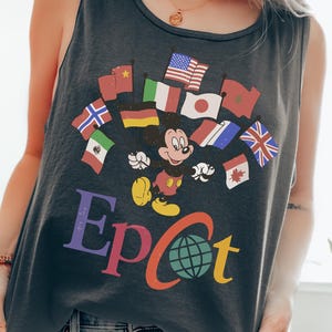 Puede incluir: Una camiseta sin mangas gris oscuro con Mickey Mouse rodeado de banderas de todo el mundo. La palabra "Epcot" está impresa en letras coloridas debajo de las banderas y Mickey Mouse. La camiseta sin mangas se combina con vaqueros azules claros.
