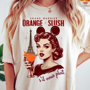 May include: A white t-shirt with a graphic of a woman with Mickey Mouse ears drinking a cocktail in front of the Eiffel Tower. The text on the shirt reads "Grand Marnier Orange Slush s'il vous plaît".