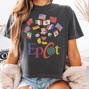 May include: Dark gray t-shirt featuring Mickey Mouse with flags and the word "Epcot" in colorful letters. The design includes flags from various countries, with a globe graphic. A casual, vintage-style tee.
