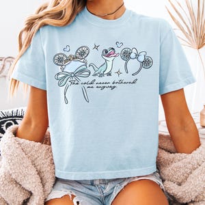 May include: Light blue t-shirt with a whimsical design featuring a bow, snowflakes, a frog, and mouse ears. The text "The cold never bothered me anyway" is written in a script font. The shirt is made of a soft material.