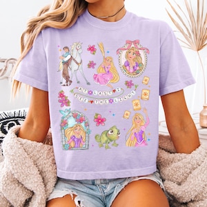 May include: A light purple t-shirt featuring a colorful graphic design with the text "AT LAST I SEE THE LIGHT". The design includes illustrations of a princess, a horse, a frog, and floating lanterns. The shirt is made of a soft material.