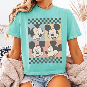 Może przedstawiać: Turkusowa koszulka z grafiką Myszki Miki w czterech kwadratach. Kwadraty mają pastelowe tła i są obramowane czarno-białym wzorem w szachownicę. Koszulka jest w swobodnym, wygodnym stylu.