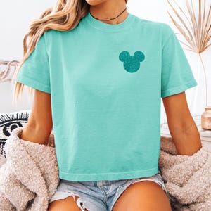 Può includere: Una maglietta turchese con un disegno glitterato della testa di Topolino sul petto sinistro. La maglietta ha maniche corte e girocollo. La persona indossa pantaloncini di jeans e un cardigan beige.