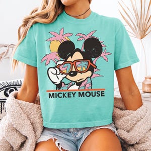 May include: A turquoise t-shirt featuring a graphic of Mickey Mouse wearing sunglasses, with a tropical sunset and palm trees. The text "MICKEY MOUSE" is printed in orange. The shirt is a relaxed fit.