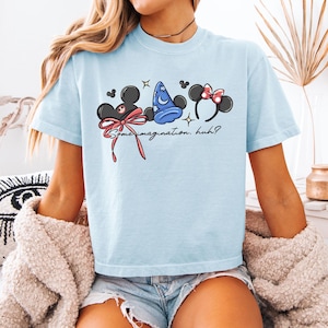 K&ouml;nnte beinhalten: Hellblaues T-Shirt mit einer Grafik von Mickey Mouse Ohren, Minnie Mouse Ohren und einem Zaubererhut. Der Text "Some imagination, huh?" ist darunter aufgedruckt. Das T-Shirt hat eine hellblaue Farbe.