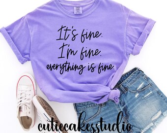 以下が含まれることがあります： 紫色のTシャツに「It's fine. I'm fine. Everything is fine.」という黒い文字が書かれています。Tシャツはデニムのショートパンツと白いスニーカーと合わせています。