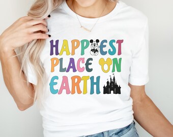 May include: A white t-shirt with the text "Happiest Place on Earth" in rainbow colors. Mickey Mouse is featured in the text and a silhouette of Cinderella's castle is to the right of the text.