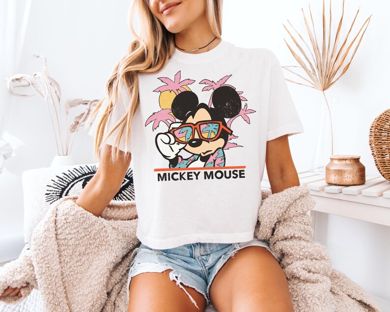 May include: White t-shirt featuring a graphic of Mickey Mouse wearing sunglasses, set against a tropical background with palm trees and a sunset. The text "MICKEY MOUSE" is printed below the graphic. The shirt is a casual, short-sleeved style.