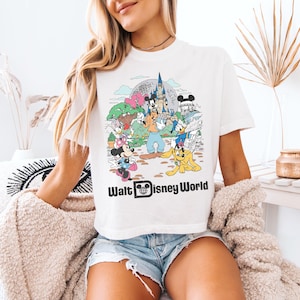 May include: White t-shirt featuring a colorful graphic of Disney characters, including Mickey Mouse, Donald Duck, and Goofy, with the text "Walt Disney World". The design includes a castle and other park elements.