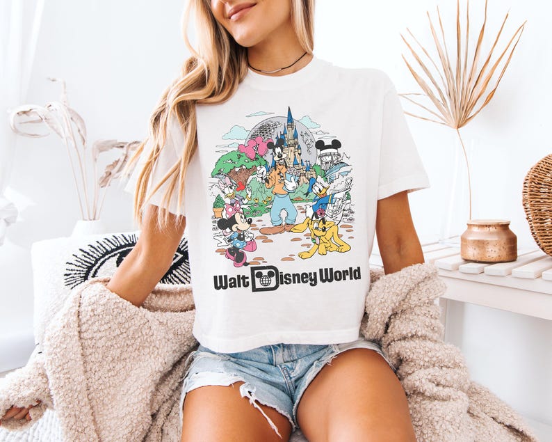 May include: White t-shirt featuring a colorful graphic of Disney characters, including Mickey Mouse, Donald Duck, and Goofy, with the text "Walt Disney World". The design includes a castle and other park elements.