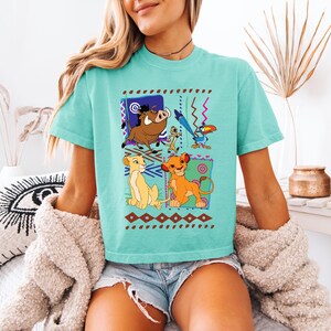 May include: A turquoise t-shirt featuring a colorful graphic of characters from The Lion King. The design includes Simba, Timon, Pumbaa, and Zazu, set against patterned backgrounds. The shirt is a comfortable, casual style.