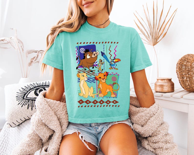 May include: A turquoise t-shirt featuring a colorful graphic of characters from The Lion King. The design includes Simba, Timon, Pumbaa, and Zazu, set against patterned backgrounds. The shirt is a comfortable, casual style.