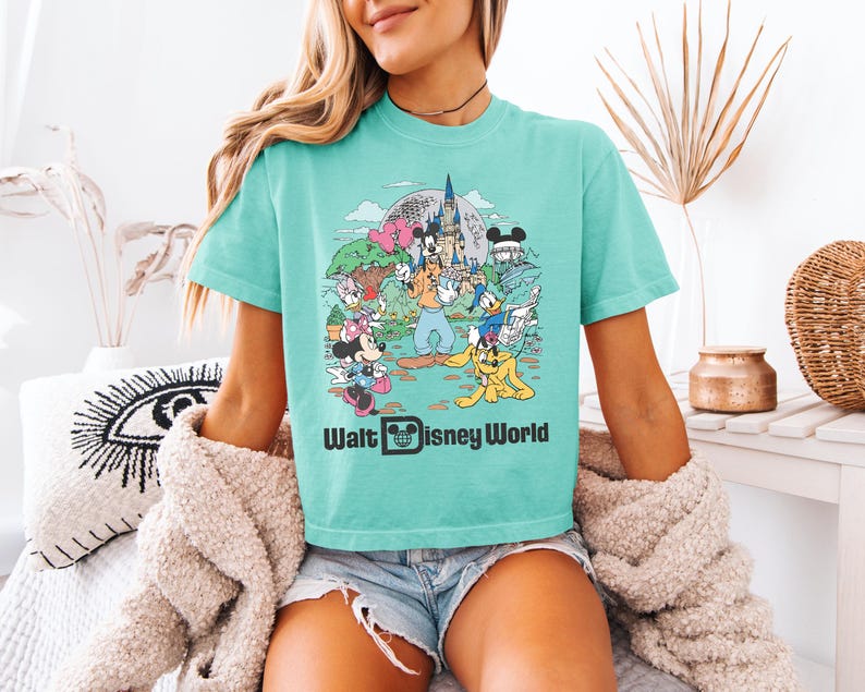 May include: A turquoise t-shirt featuring a colorful graphic of Disney characters at Walt Disney World. The design includes Mickey Mouse, Minnie Mouse, Donald Duck, and Pluto, with the text "Walt Disney World" below the image.