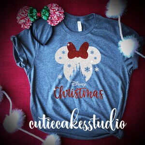 Op de afbeelding: Een grijs T-shirt met een wit en rood glitterontwerp van Mickey Mouse oren met een strik en de tekst "Disney Christmas" en de tekst "cutiecakesstudio" onderaan.