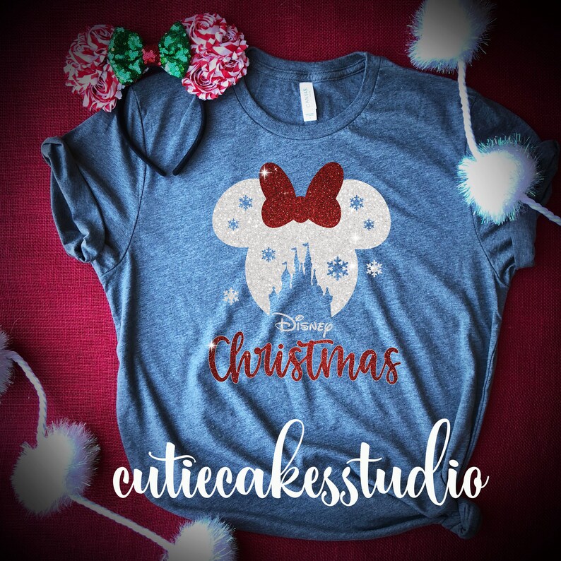 Op de afbeelding: Een grijs T-shirt met een wit en rood glitterontwerp van Mickey Mouse oren met een strik en de tekst "Disney Christmas" en de tekst "cutiecakesstudio" onderaan.