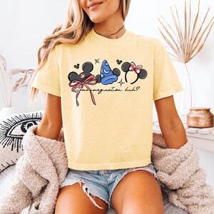K&ouml;nnte beinhalten: Hellgelbes T-Shirt mit einer Grafik von Mickey Mouse Ohren, einem blauen Zaubererhut und Minnie Mouse Ohren. Der Text "Some imagination, huh?" ist darunter aufgedruckt. Das T-Shirt ist aus weichem Material.