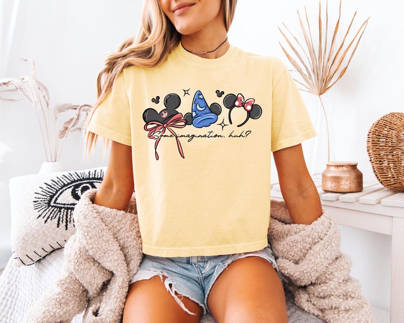K&ouml;nnte beinhalten: Hellgelbes T-Shirt mit einer Grafik von Mickey Mouse Ohren, einem blauen Zaubererhut und Minnie Mouse Ohren. Der Text "Some imagination, huh?" ist darunter aufgedruckt. Das T-Shirt ist aus weichem Material.