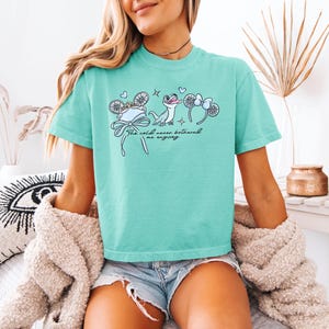 May include: A turquoise t-shirt featuring a whimsical illustration with the text "The cold never bothered me anyway." The design includes a hat, snowflakes, and a cartoon lizard with mouse ear headbands.