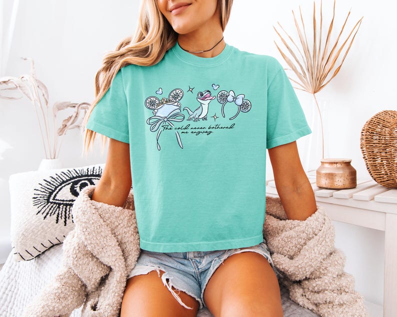 May include: A turquoise t-shirt featuring a whimsical illustration with the text "The cold never bothered me anyway." The design includes a hat, snowflakes, and a cartoon lizard with mouse ear headbands.