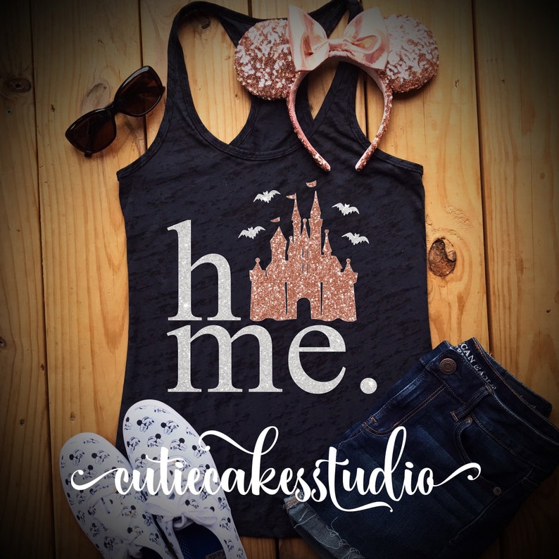 May include: A black tank top with a white glitter design that says "home" with a rose gold glitter castle and bats. The tank top is on a wooden surface with a pair of rose gold glitter mouse ears, a pair of sunglasses, and a pair of denim shorts.