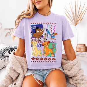 May include: A light purple t-shirt featuring a colorful graphic of characters from The Lion King. The design includes Simba, Nala, Timon, Pumbaa, and Zazu, set against a geometric background with brown and red accents.