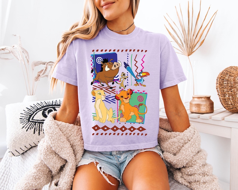 May include: A light purple t-shirt featuring a colorful graphic of characters from The Lion King. The design includes Simba, Nala, Timon, Pumbaa, and Zazu, set against a geometric background with brown and red accents.
