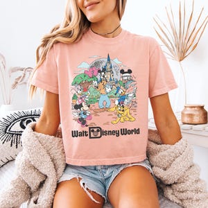 May include: A light pink t-shirt featuring a colorful graphic of Disney characters in front of a castle. The text "Walt Disney World" is printed in black below the graphic. The shirt has short sleeves and a crew neck.