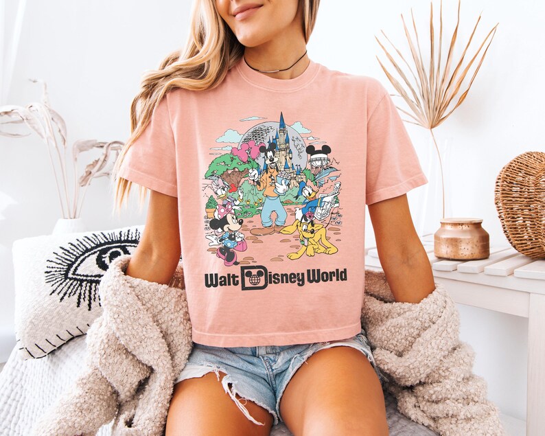 May include: A light pink t-shirt featuring a colorful graphic of Disney characters in front of a castle. The text "Walt Disney World" is printed in black below the graphic. The shirt has short sleeves and a crew neck.