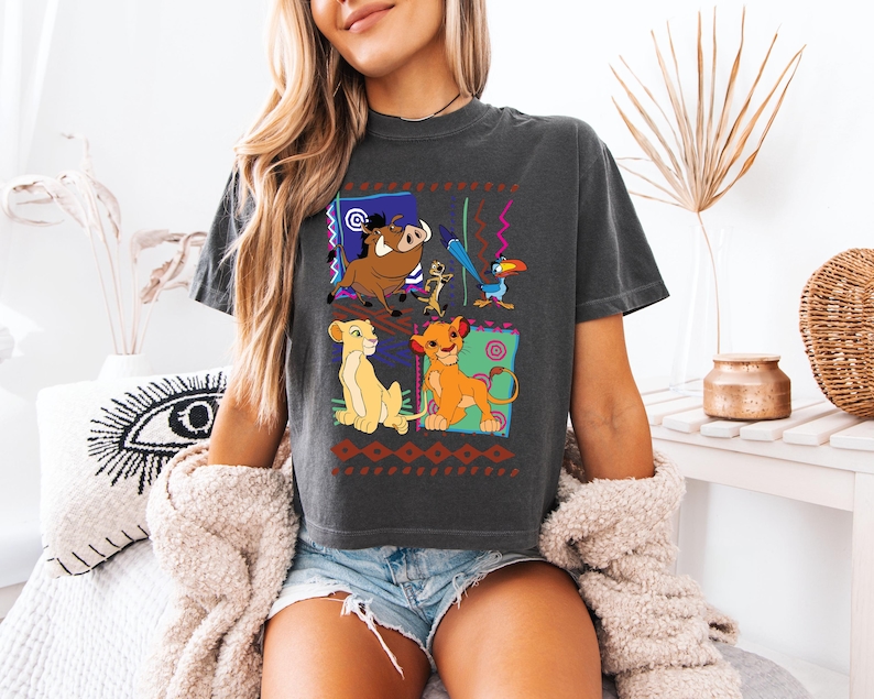 May include: Dark gray t-shirt featuring a colorful graphic of characters from The Lion King. The design includes Simba, Nala, Timon, Pumbaa, Zazu, and a toucan, set against a patterned background. The shirt is a comfortable, casual style.