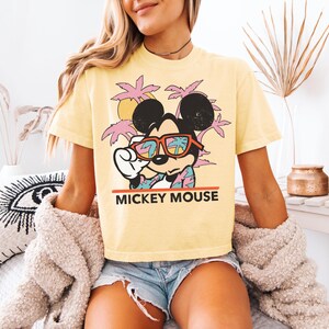 May include: Yellow t-shirt featuring Mickey Mouse wearing sunglasses with a tropical scene reflected in the lenses. The graphic includes palm trees, a sun, and the text "MICKEY MOUSE" in red.