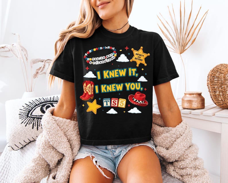 Może przedstawiać: Czarny t-shirt z kr&oacute;tkim rękawem z nadrukiem graficznym zawierającym tekst "I KNEW IT, I KNEW YOU," "YOU'VE GOT A FRIEND IN ME," "INFINITY AND BEYOND," oraz "TS5." Wz&oacute;r przedstawia bransoletkę z koralik&oacute;w, gwiazdę szeryfa, kowbojki, kapelusz i gwiazdki.