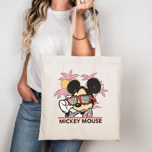 May include: Cream-colored tote bag featuring a graphic of Mickey Mouse wearing sunglasses and a Hawaiian shirt, set against a backdrop of palm trees and a sunset. The words "MICKEY MOUSE" are printed in a bold, orange font at the bottom.