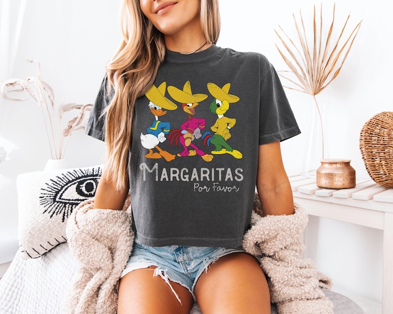 Op de afbeelding: Donkergrijs t-shirt met drie cartoonfiguren met sombrero's. Het shirt heeft de tekst "Margaritas Por Favor". De figuren zijn in heldere kleuren, met &eacute;&eacute;n in een blauw shirt, &eacute;&eacute;n in roze en &eacute;&eacute;n in groen.
