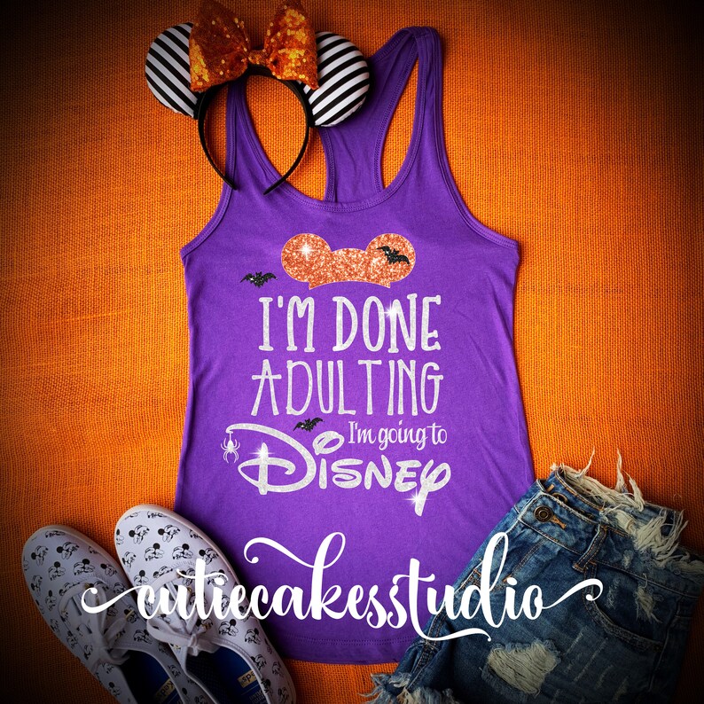 May include: Purple tank top with the text "I'm done adulting I'm going to Disney" in silver glitter with a glittery orange Mickey Mouse ear on the top. The tank top is on a orange background with a pair of white shoes with black Mickey Mouse ears on them and a pair of blue denim shorts.