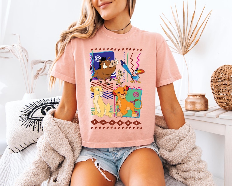 May include: A light pink t-shirt featuring a colorful graphic of characters from The Lion King. The design includes Simba, Nala, Timon, Pumbaa, and Zazu, set against a geometric background. The shirt is made of a soft material.