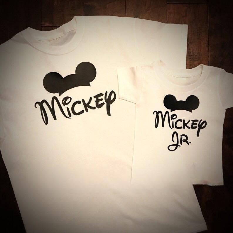 May include: Two white t-shirts with black Mickey Mouse silhouettes and the text "Mickey" and "Mickey Jr."