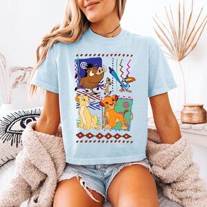 May include: Light blue t-shirt featuring a colorful graphic of characters from The Lion King. The design includes Simba, Timon, Pumbaa, and Zazu, set against a patterned background. The shirt is a casual, comfortable style.