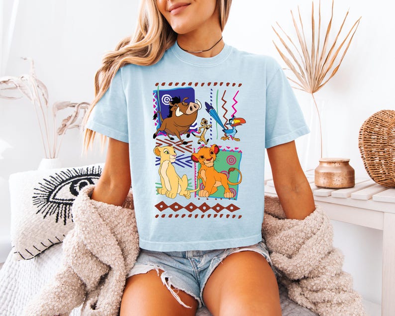 May include: Light blue t-shirt featuring a colorful graphic of characters from The Lion King. The design includes Simba, Timon, Pumbaa, and Zazu, set against a patterned background. The shirt is a casual, comfortable style.