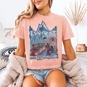 May include: A light pink t-shirt featuring a graphic of the Everest ride at Walt Disney World. The design includes the word "Everest" above a mountain scene with cartoon characters and the words "Walt Disney World" below.