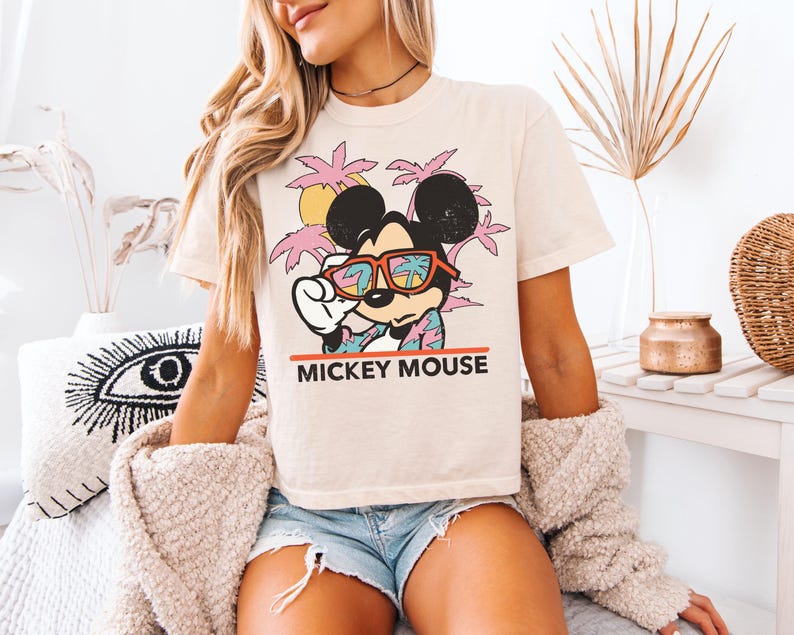 May include: Cream-colored t-shirt featuring a graphic of Mickey Mouse wearing sunglasses with a tropical scene. The text "MICKEY MOUSE" is printed in red below the image. The shirt is paired with denim shorts.