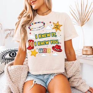 Może przedstawiać: Beżowy t-shirt z kr&oacute;tkim rękawem z nadrukiem graficznym zawierającym tekst "I KNEW IT, I KNEW YOU," "TS5" oraz "INFINITY AND BEYOND." Wz&oacute;r przedstawia bransoletkę z koralik&oacute;w, gwiazdę szeryfa, kowbojski but, kapelusz oraz klocki z literami.