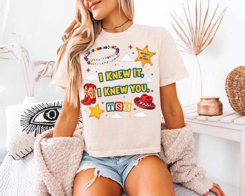Może przedstawiać: Beżowy t-shirt z kr&oacute;tkim rękawem z nadrukiem graficznym zawierającym tekst "I KNEW IT, I KNEW YOU," "TS5" oraz "INFINITY AND BEYOND." Wz&oacute;r przedstawia bransoletkę z koralik&oacute;w, gwiazdę szeryfa, kowbojski but, kapelusz oraz klocki z literami.