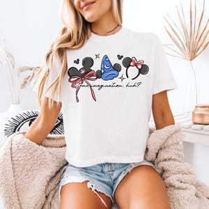 K&ouml;nnte beinhalten: Wei&szlig;es T-Shirt mit drei Cartoon-Charakter-Kopfbedeckungen: Mickey Mouse Ohren, ein blauer Zaubererhut und Minnie Mouse Ohren mit einer rosa Schleife. Der Text "Some imagination, huh?" ist unter den Designs aufgedruckt. Das Shirt wird von einer Person getragen.