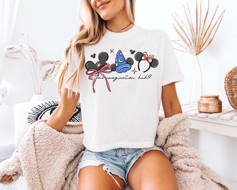 K&ouml;nnte beinhalten: Wei&szlig;es T-Shirt mit drei Cartoon-Charakter-Kopfbedeckungen: Mickey Mouse Ohren, ein blauer Zaubererhut und Minnie Mouse Ohren mit einer rosa Schleife. Der Text "Some imagination, huh?" ist unter den Designs aufgedruckt. Das Shirt wird von einer Person getragen.